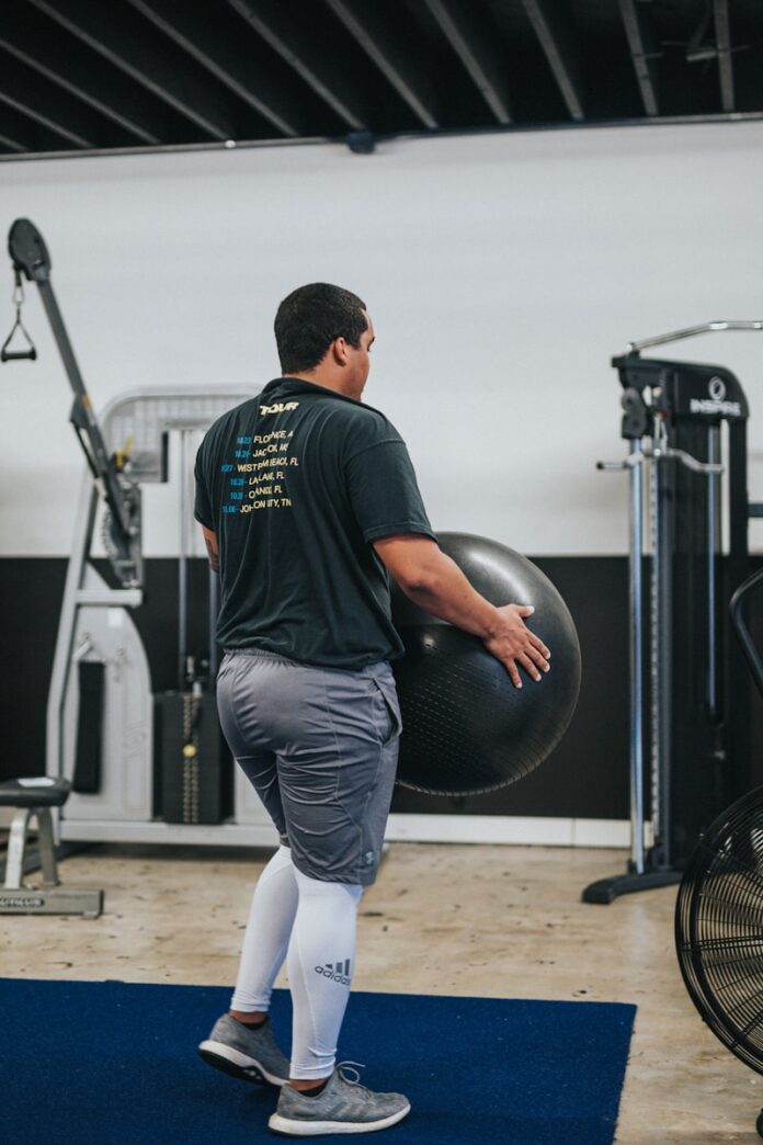 Photo by Andrew Valdivia man in black t-shirt and blue denim jeans holding black exercise ball